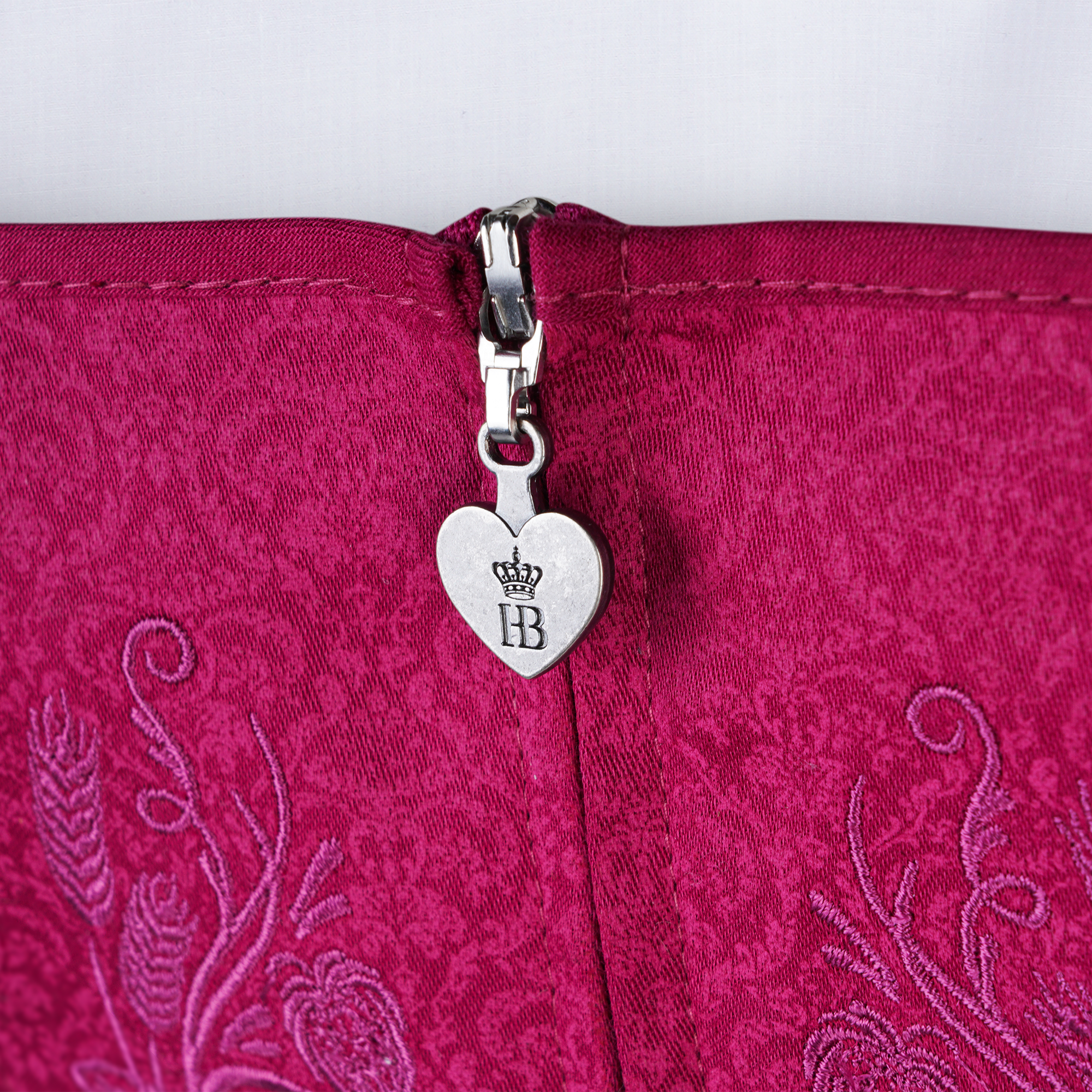 Nahaufnahme des Reißverschlusses eines pinken Dirndls mit floraler Struktur. Der Zipper ist mit einem herzförmigen Anhänger verziert, auf dem das „HB“-Logo mit Krone eingraviert ist.