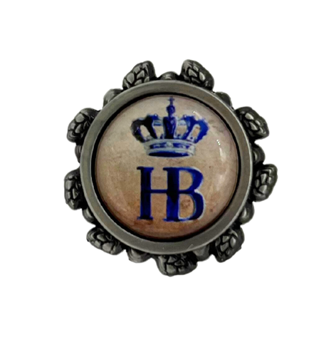 Runder Metall-Pin mit beigem Hintergrund und dekorativem Rand. Im Zentrum befindet sich das blaue Monogramm „HB“ mit einer stilisierten blauen Krone darüber – ein Symbol für das Hofbräuhaus. Der Rand ist mit feinen Ornamenten verziert, die an Hopfenranken