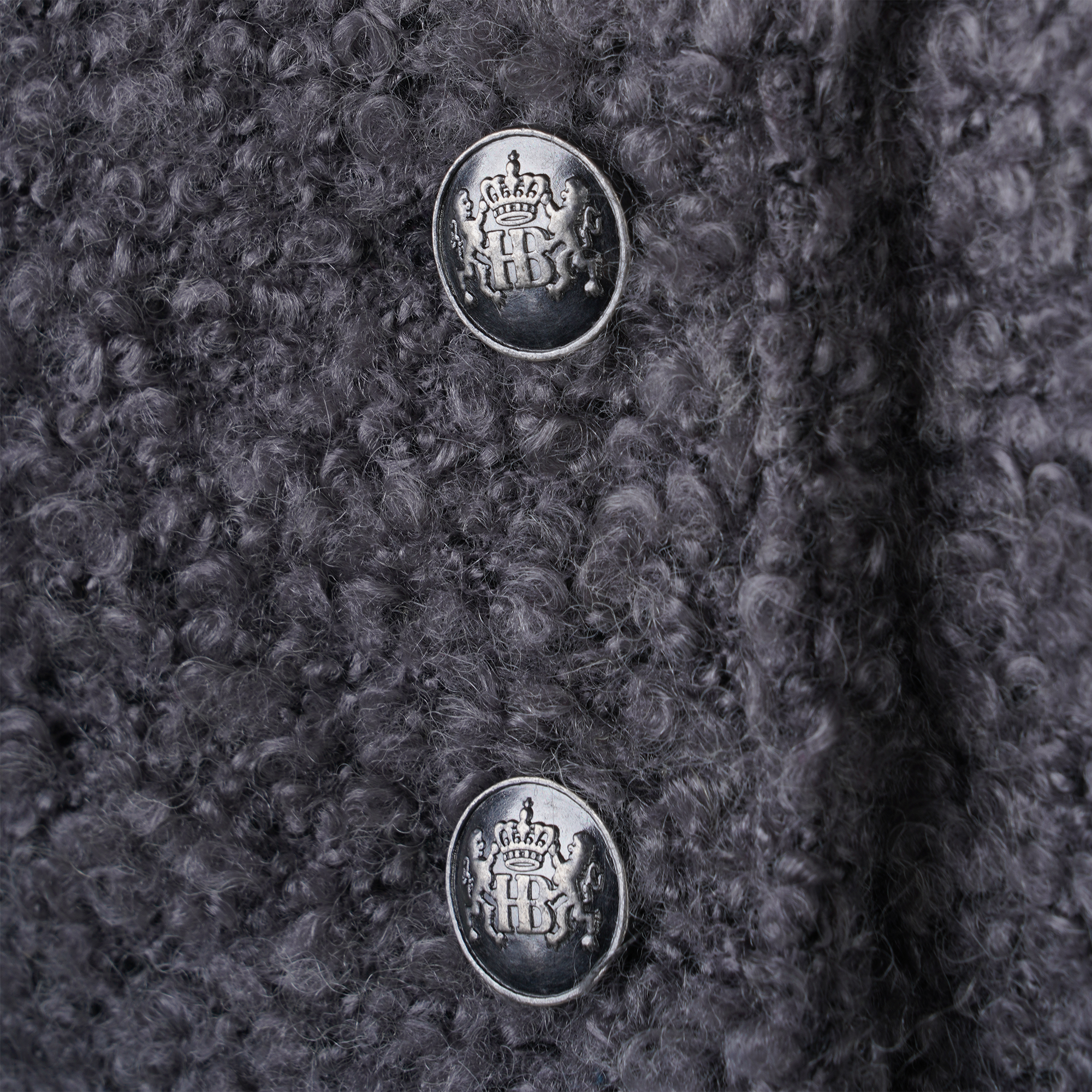 Nahaufnahme von zwei silberfarbenen Knöpfen auf dunkelgrauem Strickstoff, jeweils mit „HB“-Logo und Krone verziert.