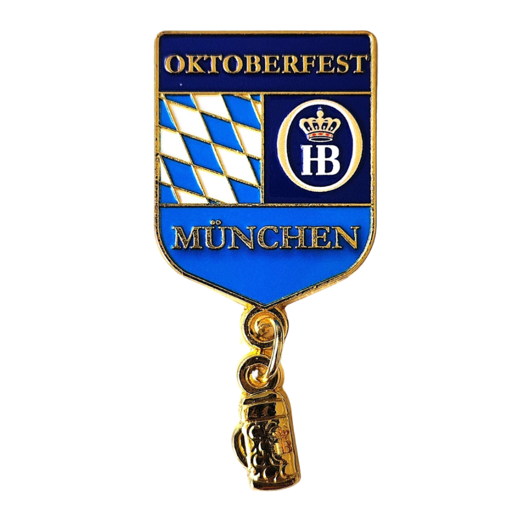 „Ansteckpin mit blau-weißem Rautenmuster auf der linken Seite, das an die bayerische Flagge erinnert. Rechts daneben befindet sich ein Kreis mit den weißen Buchstaben ‚HB‘ auf blauem Hintergrund. Darüber steht der Schriftzug ‚OKTOBERFEST‘, darunter ‚MÜNCH