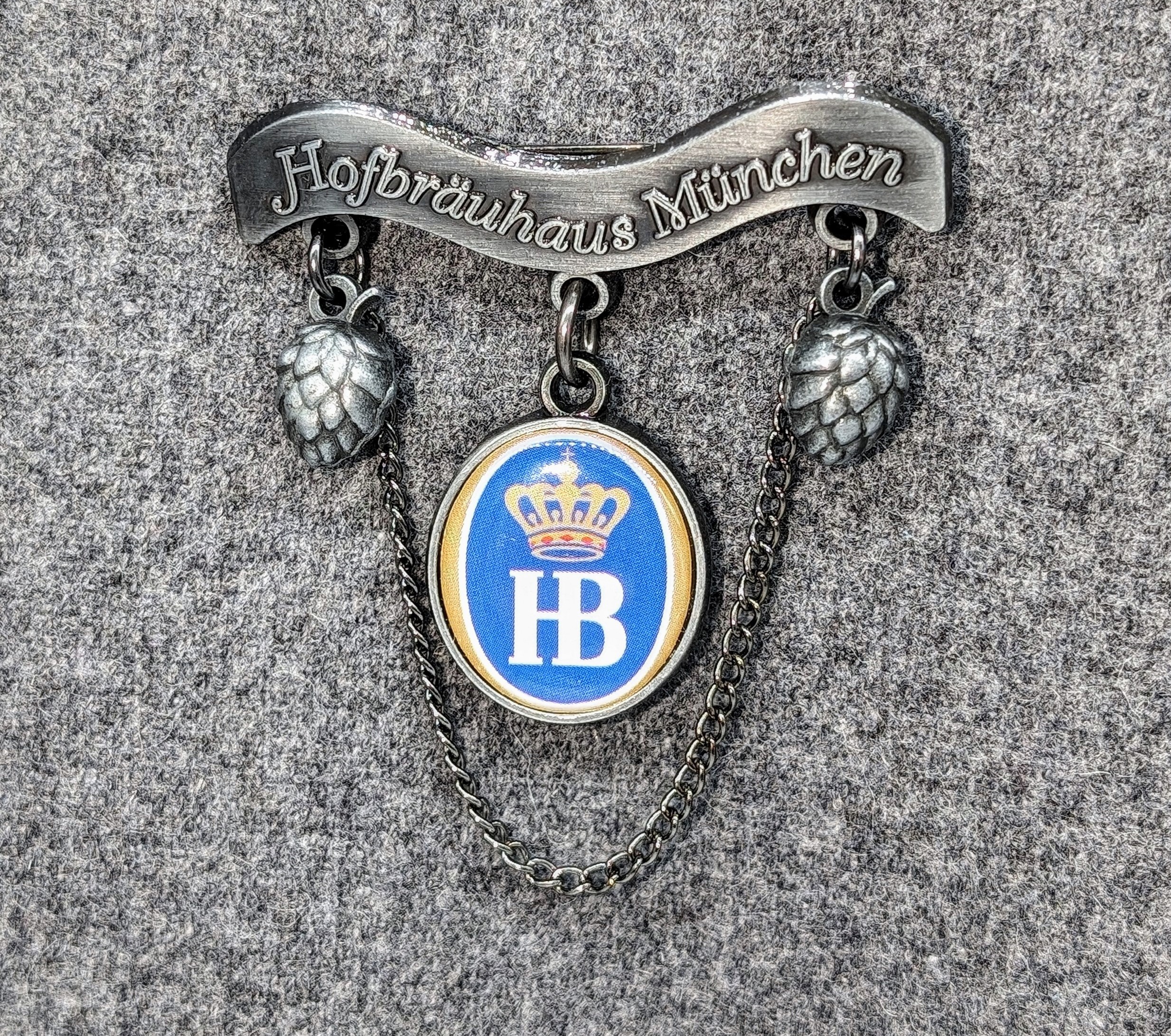 Dekorativer Metall-Anstecker mit einem oberen Banner, auf dem „Hofbräuhaus München“ steht. Darunter hängen zwei stilisierte Hopfendolden seitlich herab. In der Mitte befindet sich ein rundes Emblem mit blauem Hintergrund, goldener Krone und den weißen Buc