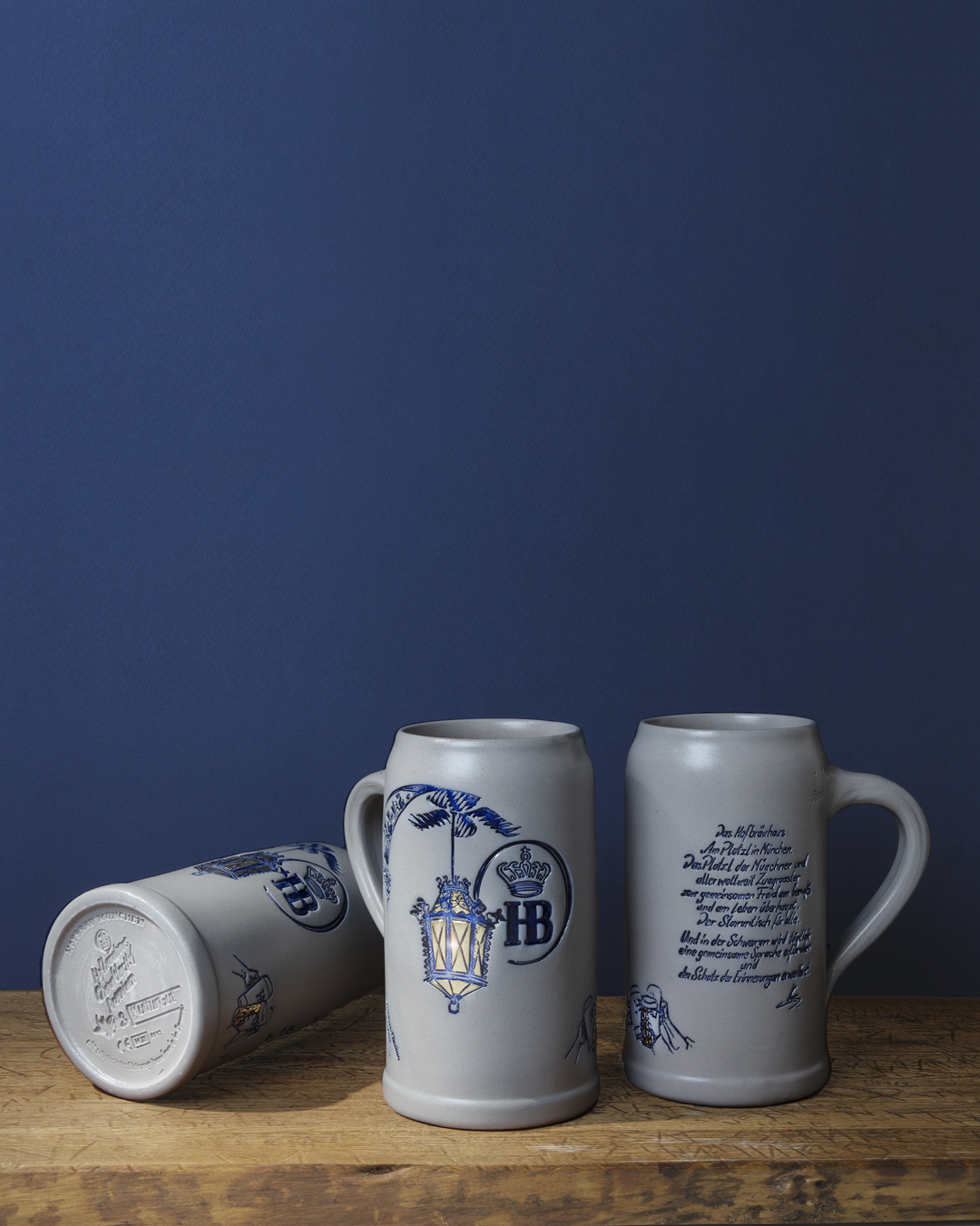 „Drei Keramik-Bierkrüge mit Henkel auf Holzfläche vor blauem Hintergrund: links ein liegender Krug mit sichtbarem Boden und Aufdruck, in der Mitte ein stehender Krug mit ‚HB‘-Emblem und floralen Verzierungen, rechts ein Krug mit deutschem Schriftzug.“