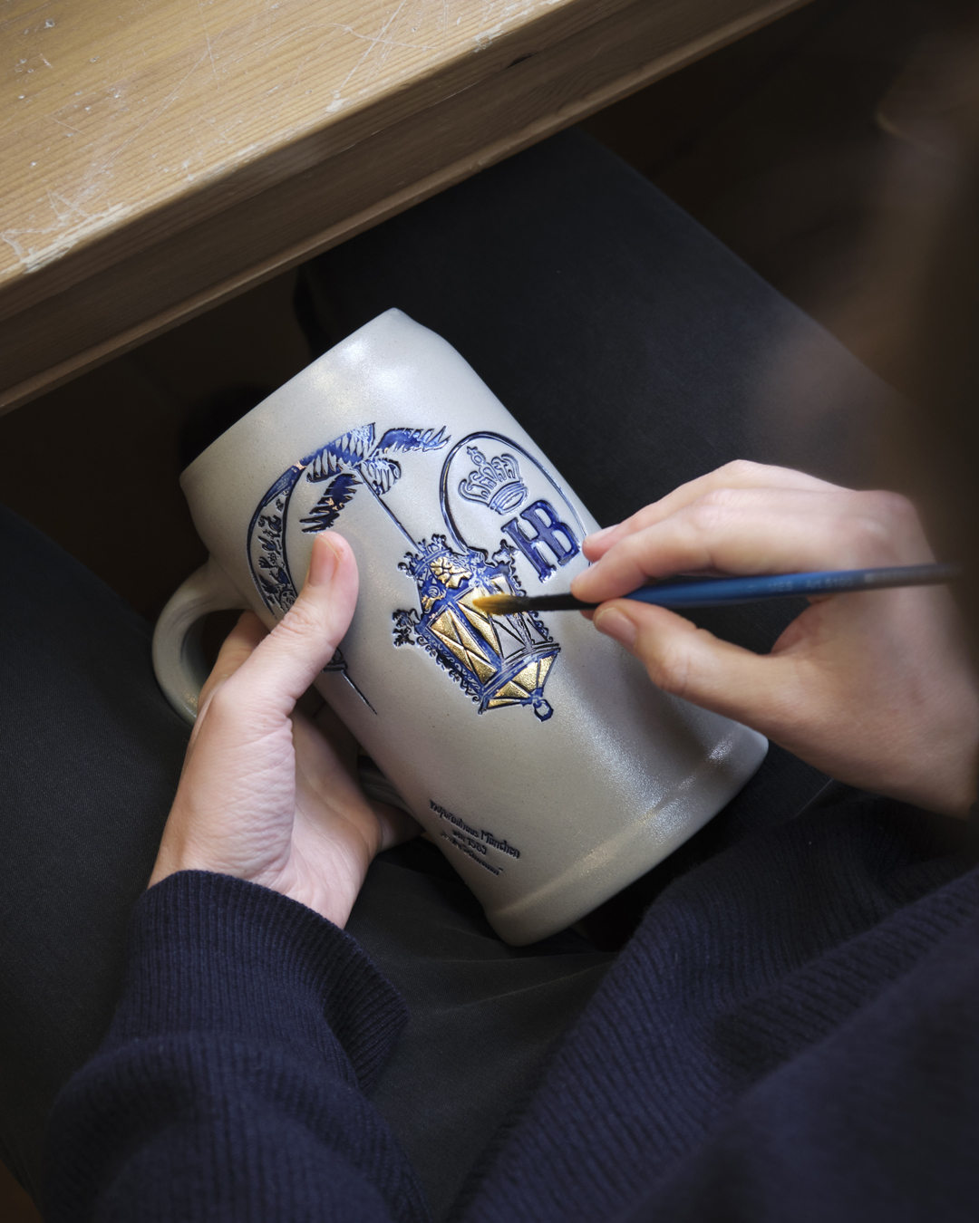 „Person bemalt einen Bierkrug mit Hofbräuhaus-München-Logo, bestehend aus Krone und HB-Schriftzug, Fokus auf Hände und Keramik.“